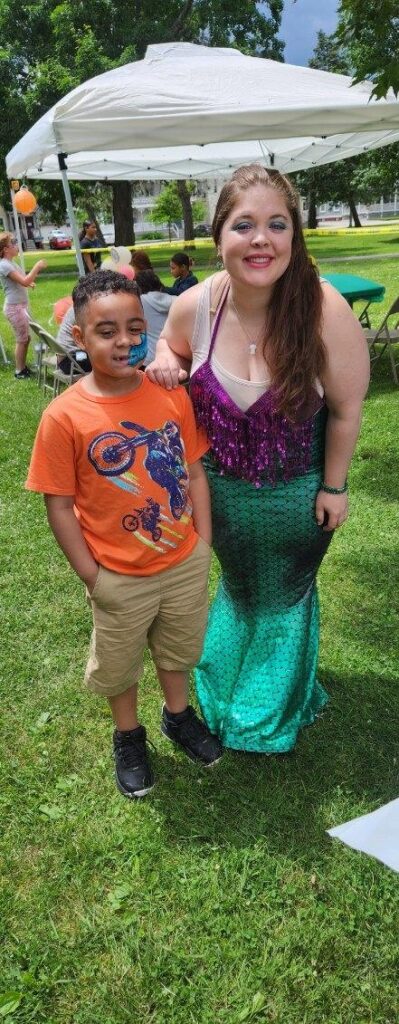 Alexis as a mermaid posing with a boy who has his face painted under a tent at an outdoor festival | Magicians for Kids Party